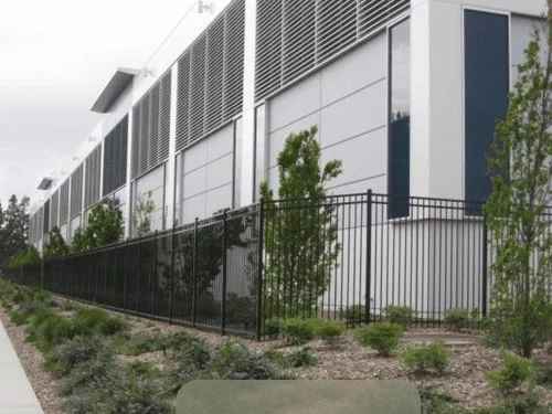 Modern industrial building with large vents and blue panels, bordered by a black metal fence and landscaped with small trees and shrubs, under an overcast sky.