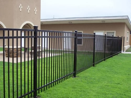 A black metal fence borders a lush, green lawn beside beige buildings with arched windows. The scene is calm and orderly under a cloudy sky.