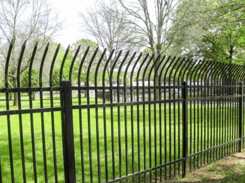 Black metal fence with pointed bars curves inward, enclosing a lush green park. Trees with sparse leaves stand against a bright, cloudy sky.