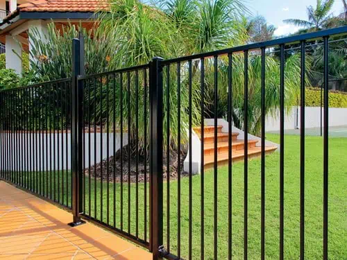 A black metal fence borders a lush green lawn in front of a house. Steps lead up to the entrance, surrounded by tropical plants and palm trees.
