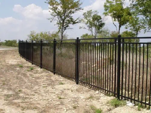A long black metal fence runs alongside a dirt path, bordered by grass and sparse trees under a partly cloudy sky, conveying a sense of openness.
