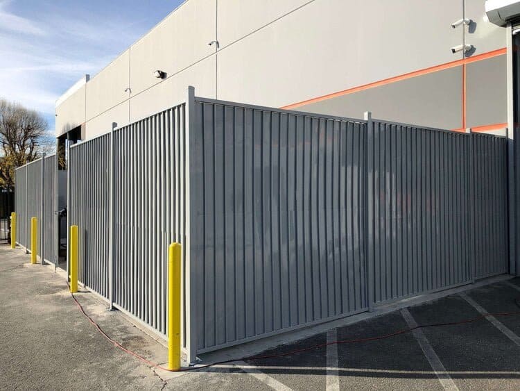 Gray metal fence enclosure with vertical slats beside a commercial building. Bright yellow bollards stand nearby, under a clear blue sky.