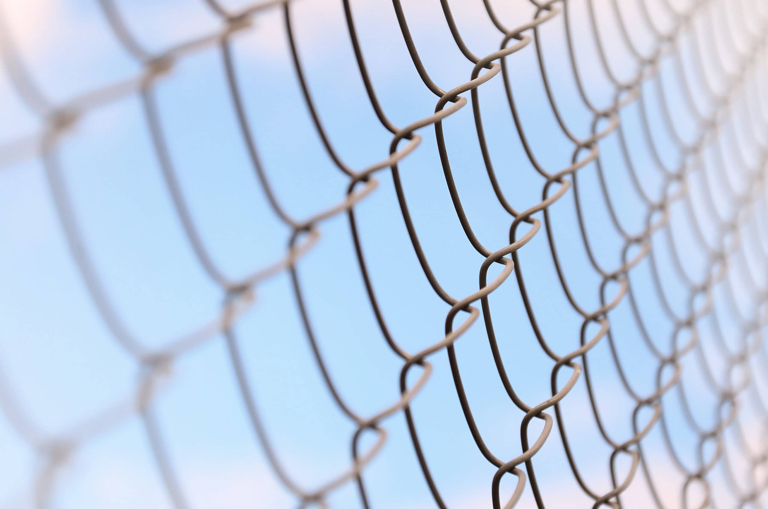 Close-up of a chain-link fence with a focus on its metal loops against a softly blurred blue sky, evoking a sense of openness and confinement.